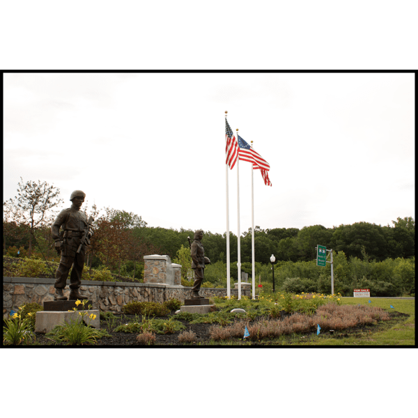 photo of garden setting and rock wall, flags, and trees behind with two bronze-colored sculptures of soldiers