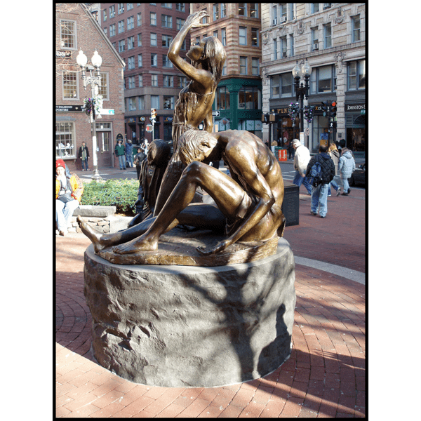 photo of bronze-colored sculpture of male, female, and child in movement on round stone base in hardscaped plaza with buildings and streets surrounding it
