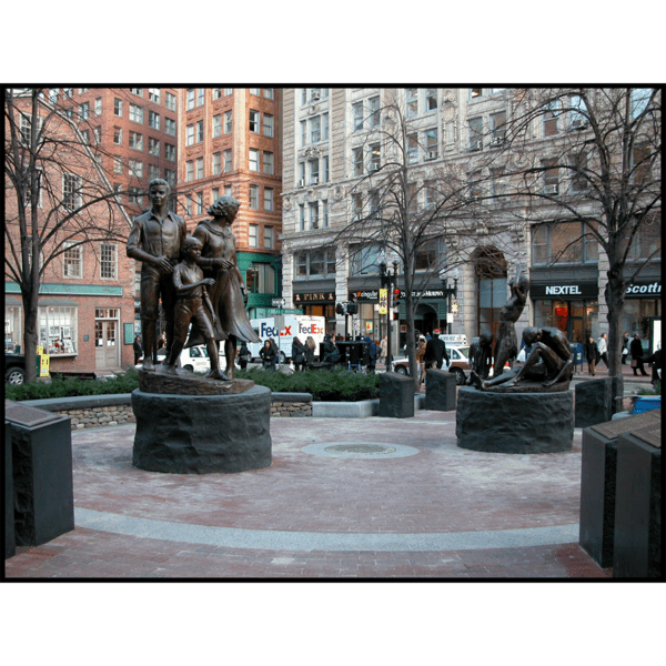 photo of two bronze-colored sculptures of a male, female, and child in movement on round stone bases in hardscaped plaza with buildings and streets surrounding it