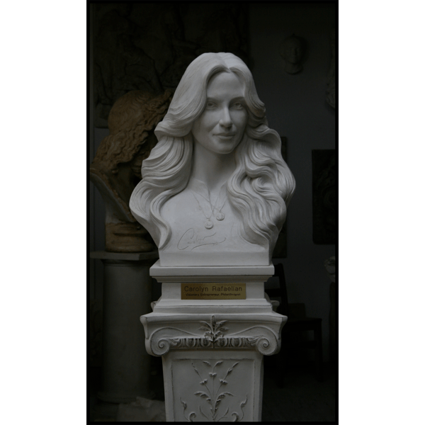 photo of white-colored sculpture bust of Carolyn Rafaelian on white square base with small gold-colored plaque on ornate white pedestal against a black background