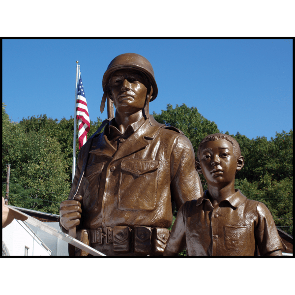 photo of bronze-colored sculpture of American male soldier and Korean boy with flag and trees behind