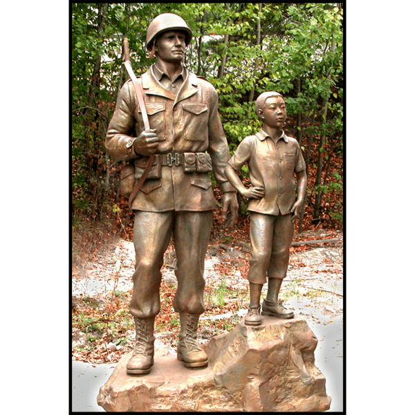 photo of bronze-colored sculpture of American male soldier and Korean boy posed on natural-looking rock with trees behind