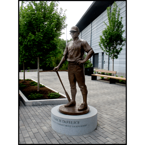 photo of bronze statue of man in baseball uniform holding bat in hand while it rests on ground on a circular granite base in a plaza with building and trees behind