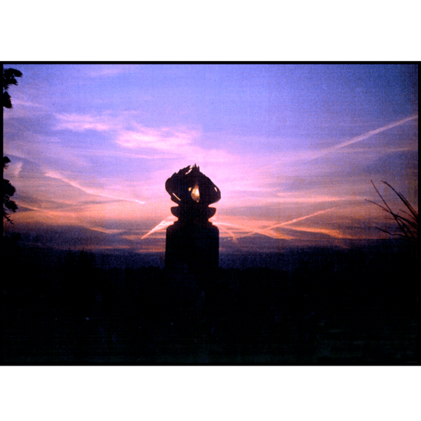 photo of purple and pink sunset and silhouette of bronze sculpture of flame on black granite base with surrounding trees