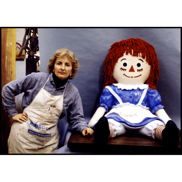photo of sculpture of Raggedy Ann doll with polychrome finish on gray background with sculptor Kathleen Shure standing next to it