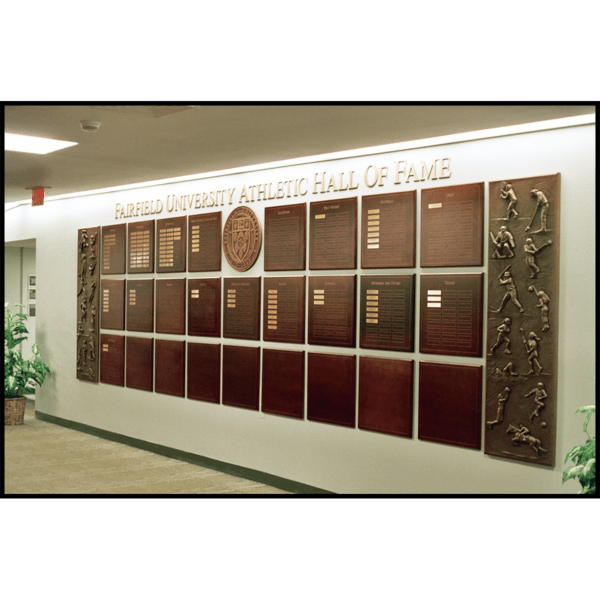 photo of white interior wall with rows of bronze-colored plaques with names on small gold-colored plaques with two large bronze-colored reliefs of various athletes