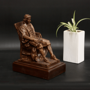photo of bronze-colored sculpture of John Harvard seated in a chair atop a wood base beside a white vase with a plant