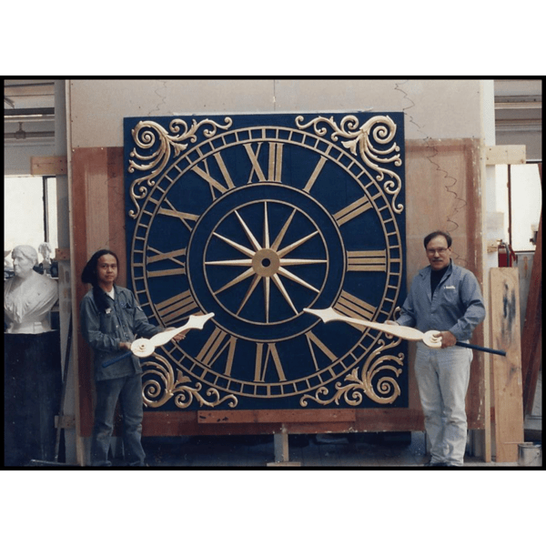 photo of blue and gold clock in studio with sculptor Robert Shure and assistant standing beside it holding the gold clock hands