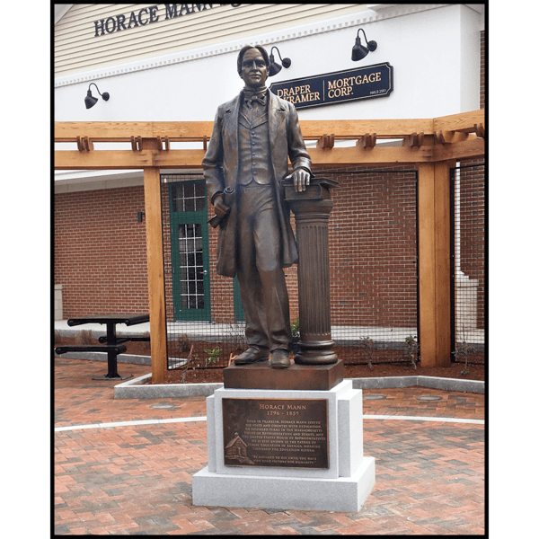 photo of bronze-colored statue of Horace Mann holding scroll and standing beside column with books, atop granite pedestal with bronze-colored plaque in a hardscaped plaza with building behind