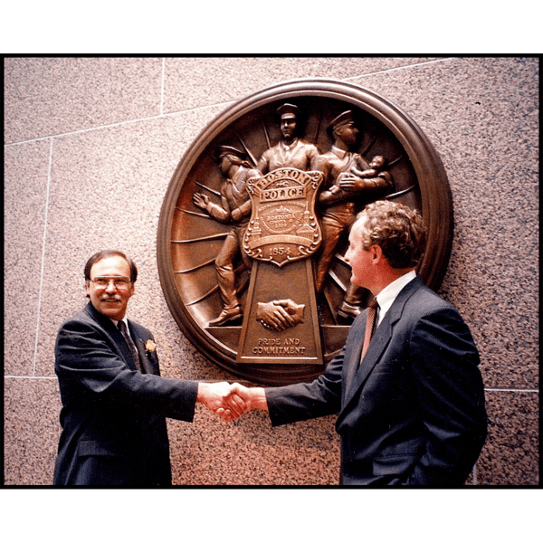 photo of sculptor Robert Shure shaking hands with Boston official at dedication ceremony for Police and Fire department plaques, standing in front of large, bronze-colored police plaque mounted on stone wall