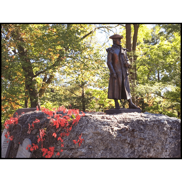 photo of bronze statue of Joseph Warren in uniform holding sword hilt at his side atop a large puddingstone with red leaves on left side