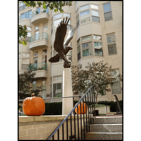 photo of bronze sculpture of eagle perched on ball with open wings atop tall pedestal at the top of a staircase with two pumpkins in front of an off-white building and pumpkins