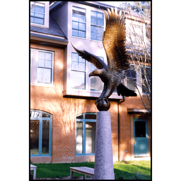 photo of bronze sculpture of eagle perched on ball with open wings atop tall pedestal in front of a brick building