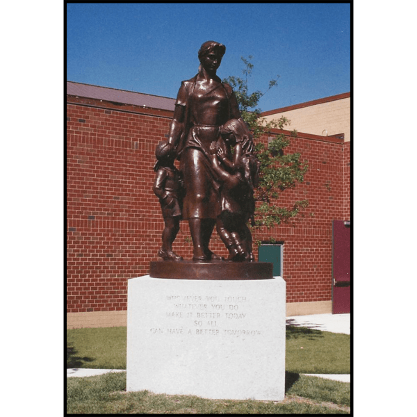 photo of bronze-colored sculpture of female teacher with three children atop granite pedestal on grass in front of brick building