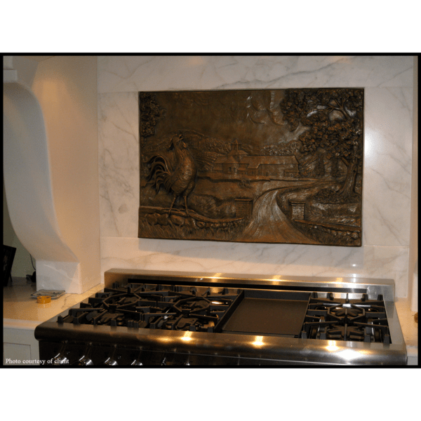 photo of bronze-colored relief plaque of farmhouse with rooster and tree in foreground mounted on tiled wall above kitchen stove