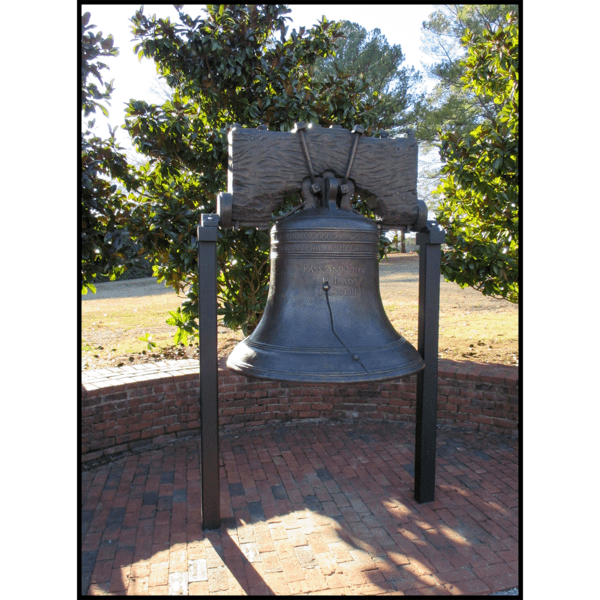 photo of bronze-colored Liberty Bell with yoke and held up with two posts in brick plaza with grass and trees in background