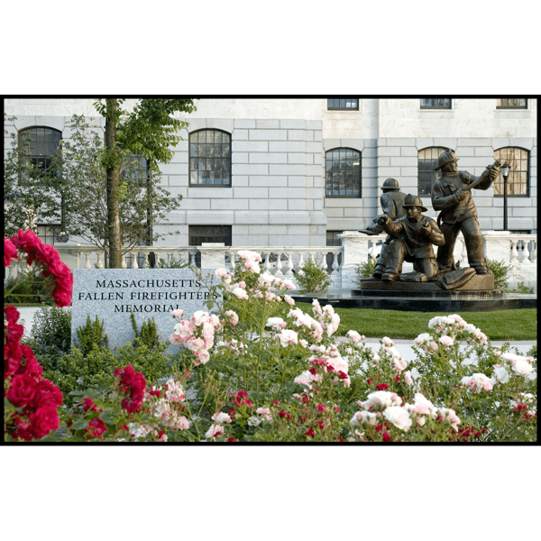 photo of bronze sculpture of three firefighters in action stances on a stone base in front of white stone building with grass and flowers in foreground