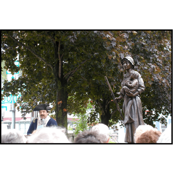 photo of person in costume and a crowd in front of bronze statue of Molly Stark holding child and musket during dedication ceremony with surrounding trees