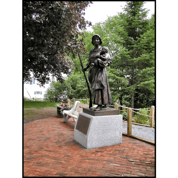photo of bronze statue of Molly Stark holding child and musket atop stone base with plaque on a brick plaza with surrounding trees