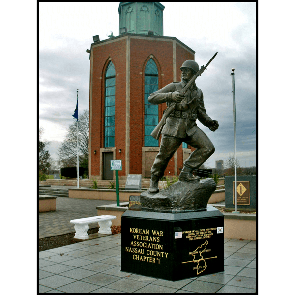 photo of bronze-colored sculpture of soldier holding rifle in action on incline on stone base in plaza and building behind