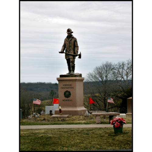 photo of bronze statue of standing firefighter holding an ax atop a tall stone base in a cemetery