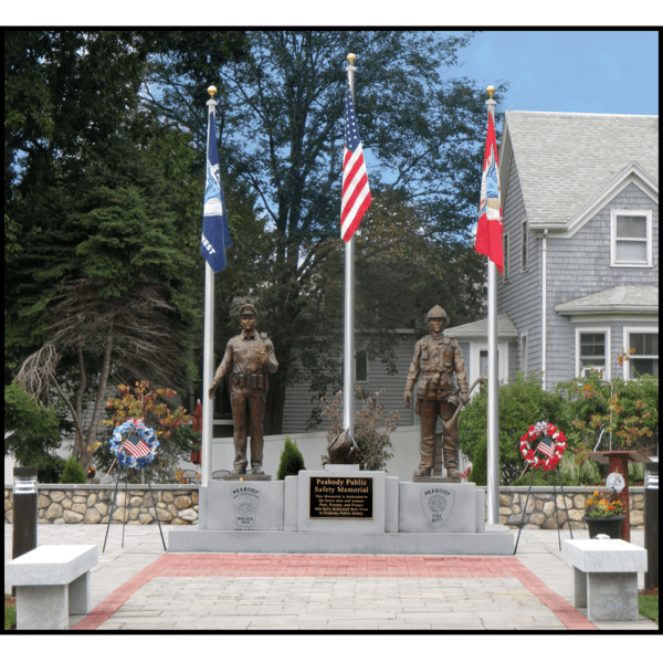 photo of outdoor memorial of bronze sculptures of a firefighter, a police officer, and an eternal flame on a large stone base with etchings and a bronze plaque in a paved site with three flags behind and trees and building behind