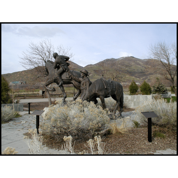 photo of bronze sculpture of galloping horse with rider and a man and another horse drinking water in a plaza with a mountain range behind