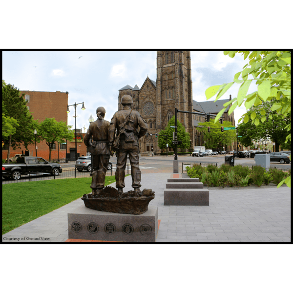 photo from the back of bronze sculpture of two soldiers standing next to one another on low base with benches in middle ground and church in background
