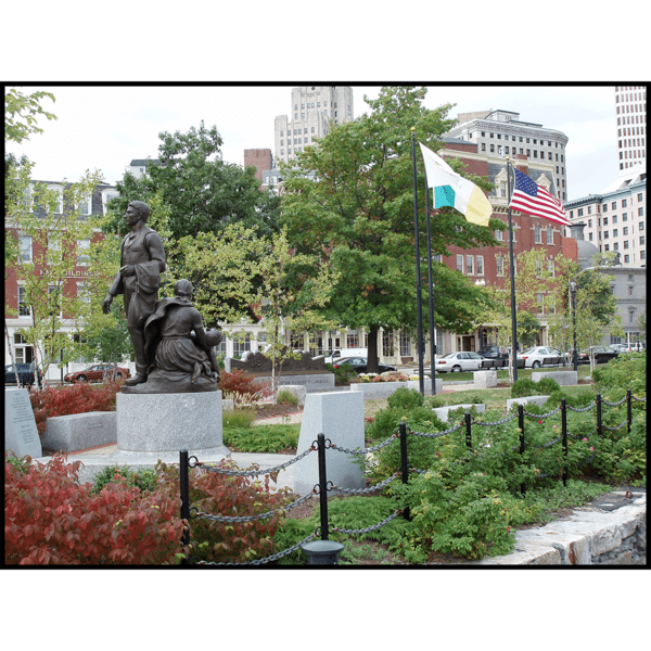 photo of bronze figural group sculpture on round stone base surrounded by pathway, fence, flags, shrubs, and trees
