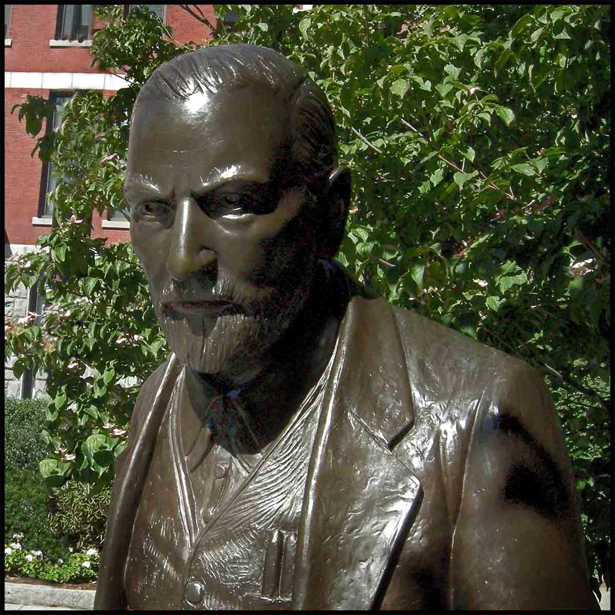 photo closeup of face of bronze sculpture of Sigmund Freud sitting on a stone bench in a plaza with trees behind