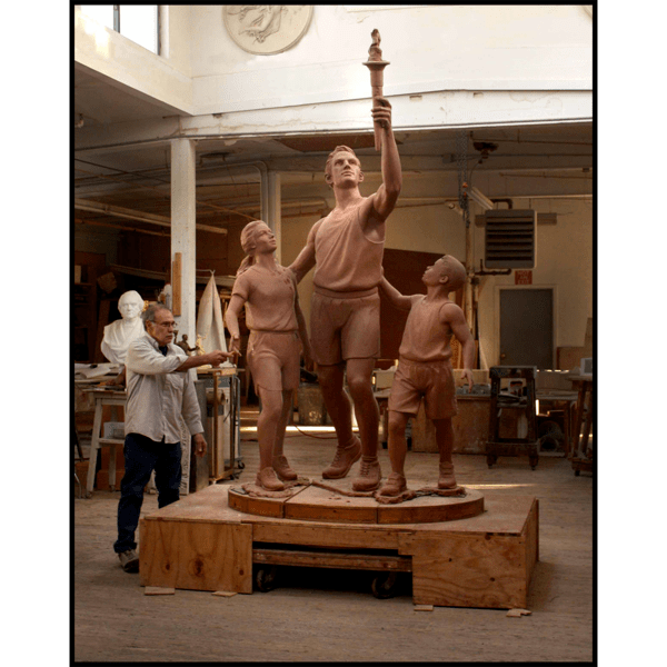 photo of clay model of sculpture of man holding up Olympic torch with a boy and girl at his sides, with artist Robert Shure in his studio