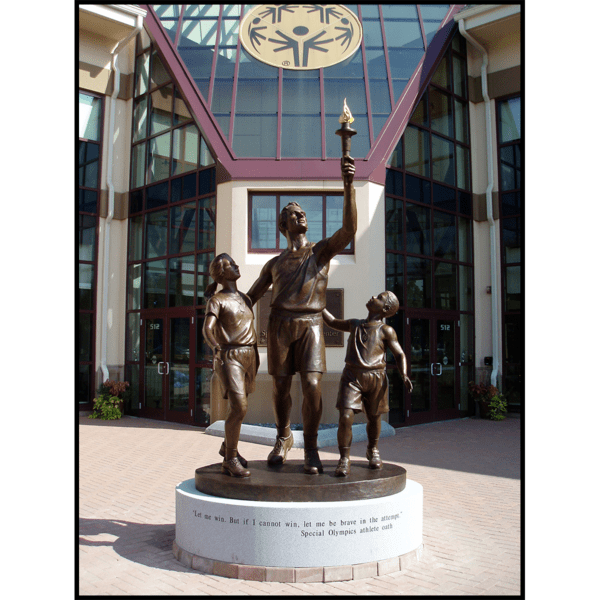 photo of bronze sculpture of man holding up Olympic torch with a boy and girl at his sides, placed on round stone base in plaza in front of building