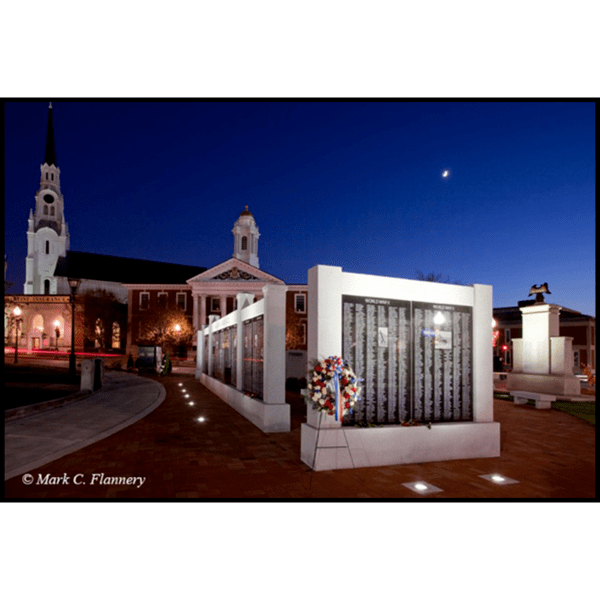 photo of black and gray granite monument in form of a wall on brick hardscaping in city green with buildings and trees behind at night