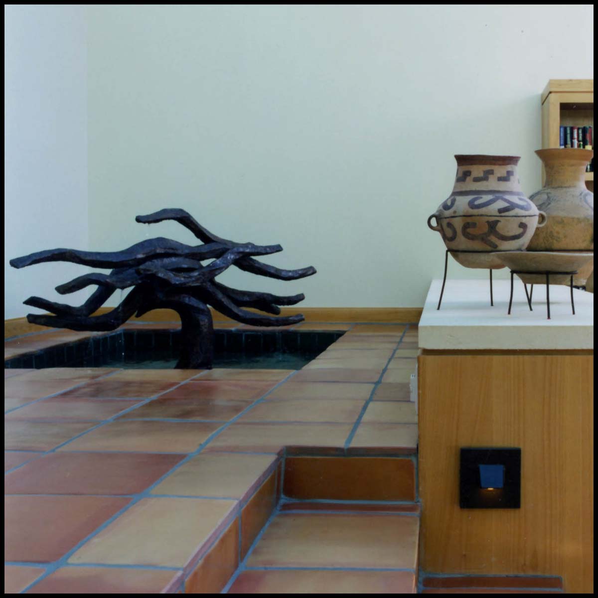 photo of bronze-colored small indoor fountain with flowing shapes, in white-walled room with tiled floor, tiled steps, and wooden shelves and bookcases