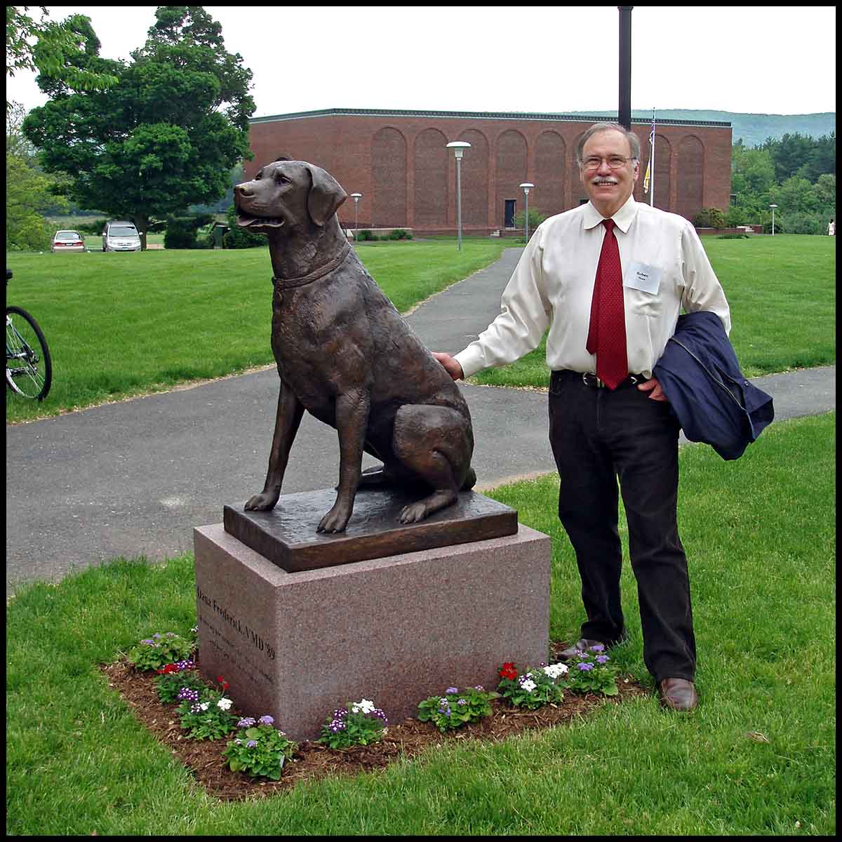 photo of bronze-colored sculpture of black labrador on stone base on landscaped campus with sculptor Robert Shure beside it