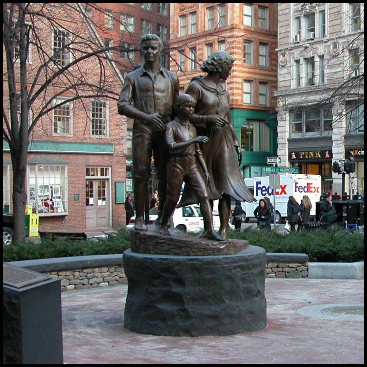photo of bronze-colored sculpture of male, female, and child in movement on round stone base in hardscaped plaza with buildings and streets surrounding it