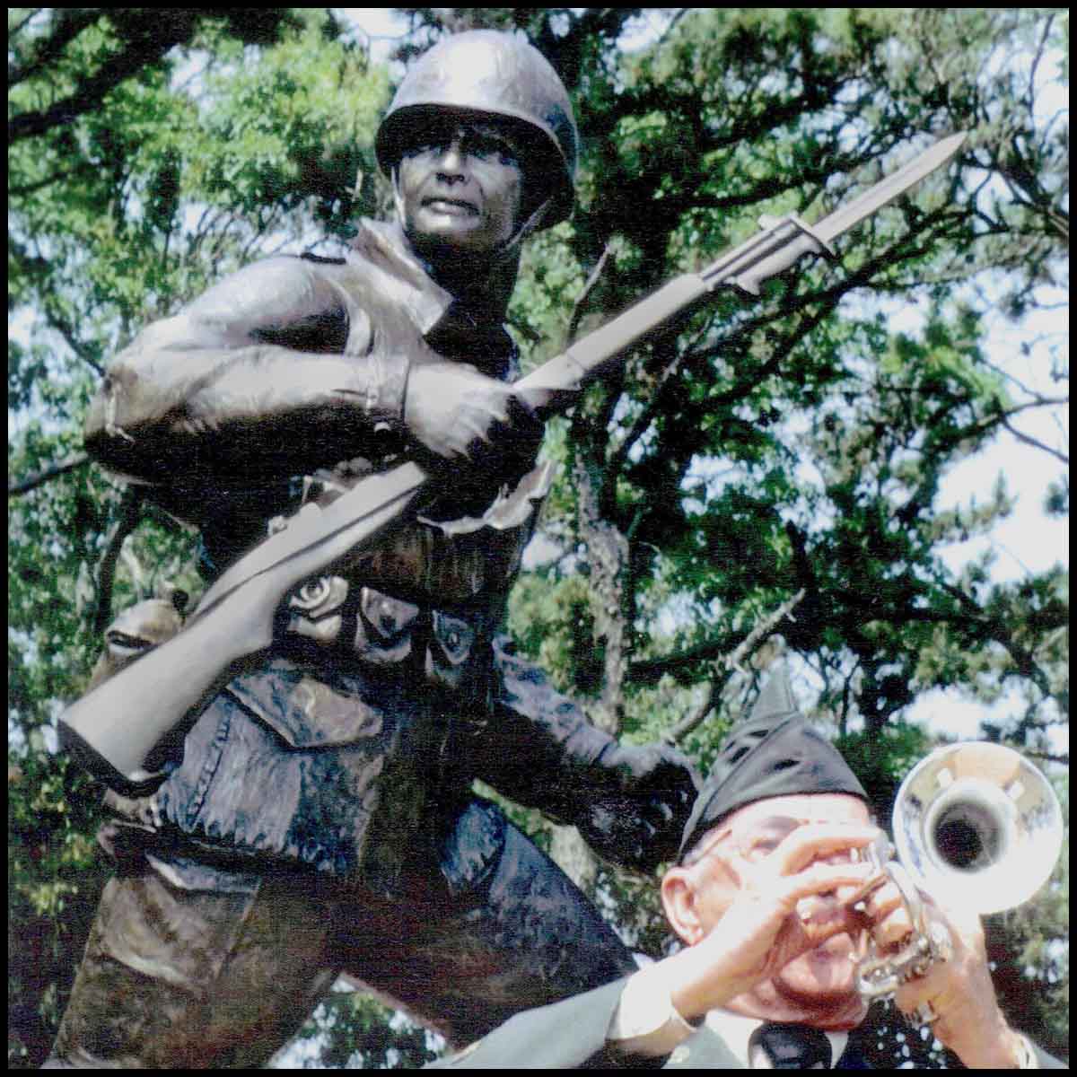 closeup photo of bronze-colored sculpture of soldier holding rifle in action on incline with trees behind and veteran in dress uniform in front playing trumpet
