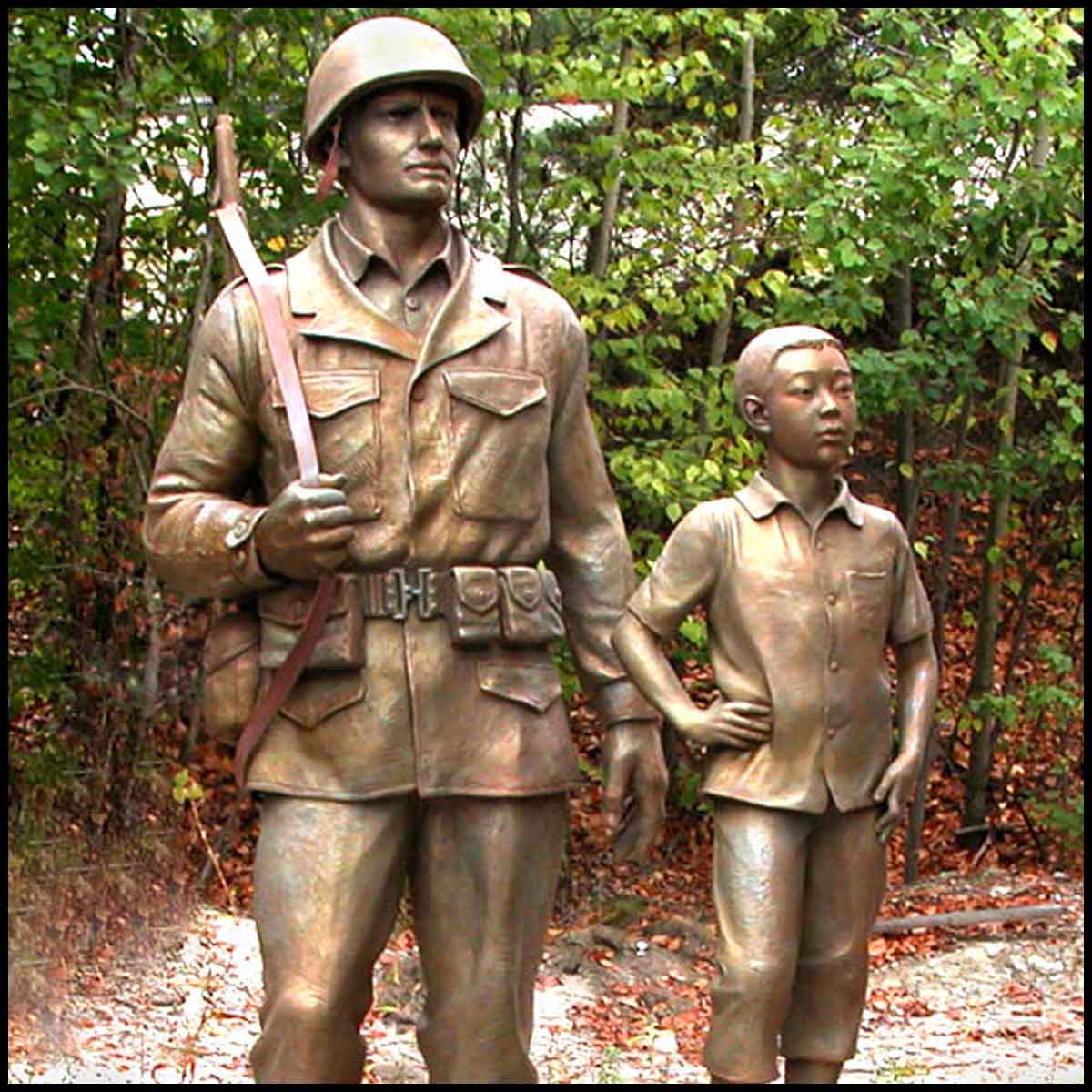 photo of bronze-colored sculpture of American male soldier and Korean boy with trees behind