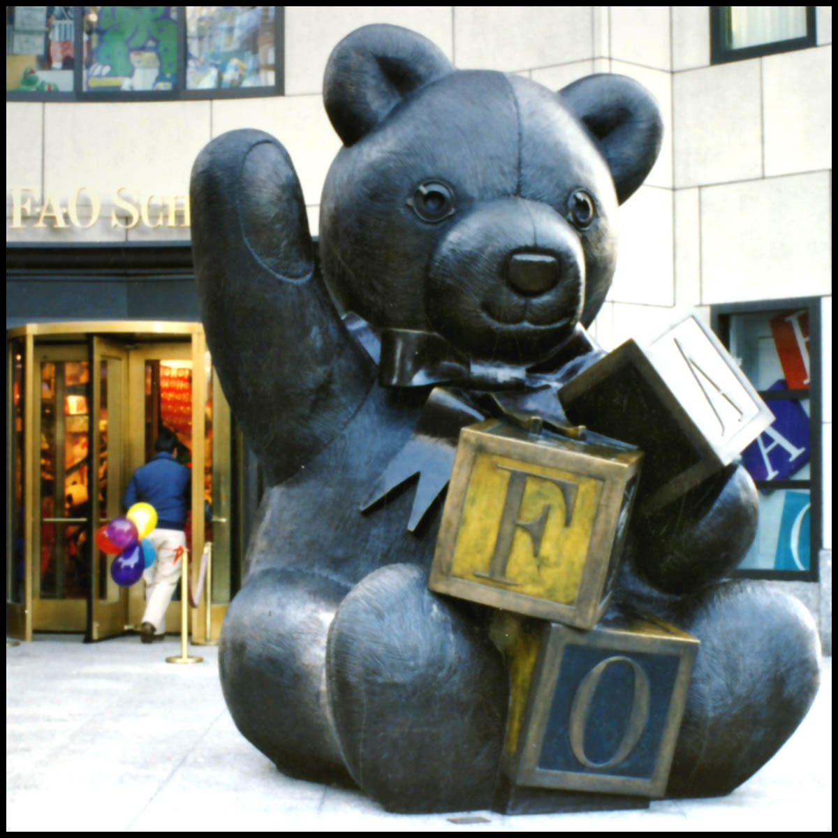 photo of bronze teddy bear with arm raised and holding blocks in front of city store