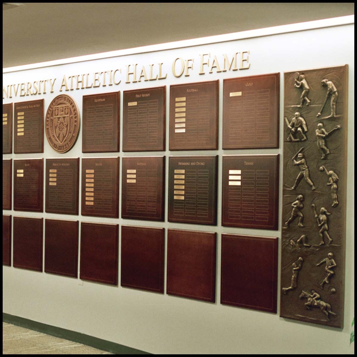 photo of white interior wall with rows of bronze-colored plaques with names on small gold-colored plaques with large bronze-colored relief of various athletes