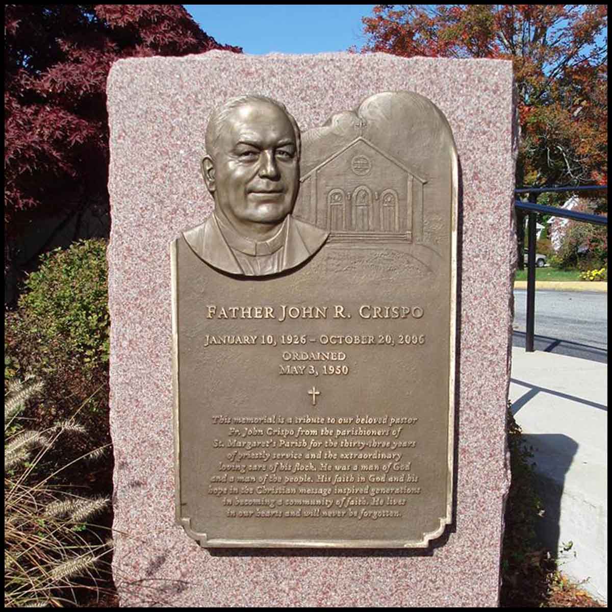 photo of bronze-colored plaque with portrait of John Crispo mounted on granite in landscaped area with walkway with railing and trees behind