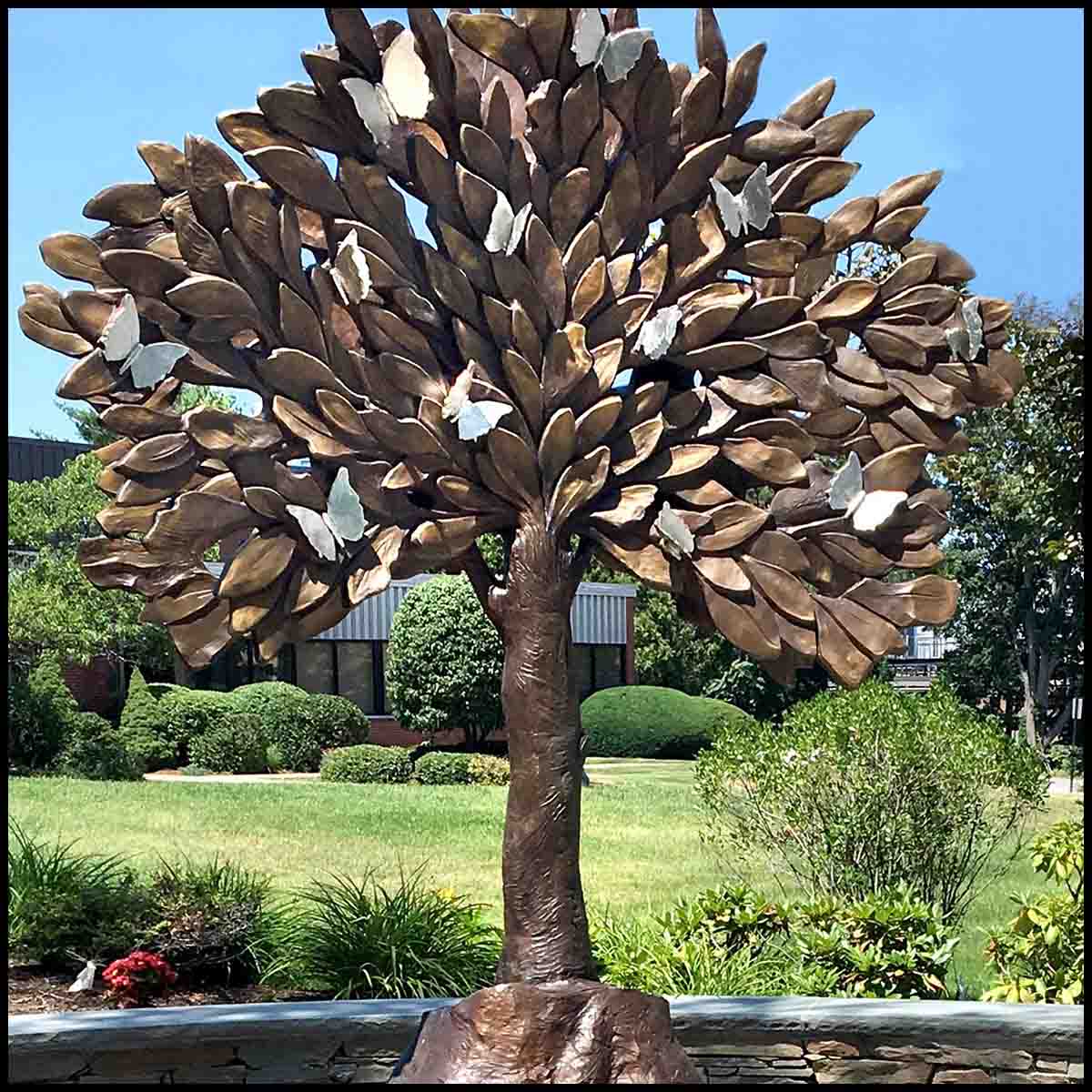 closeup photo of bronze-colored tree sculpture on granite base in hardscaped plaza surrounded by landscaping with building in background