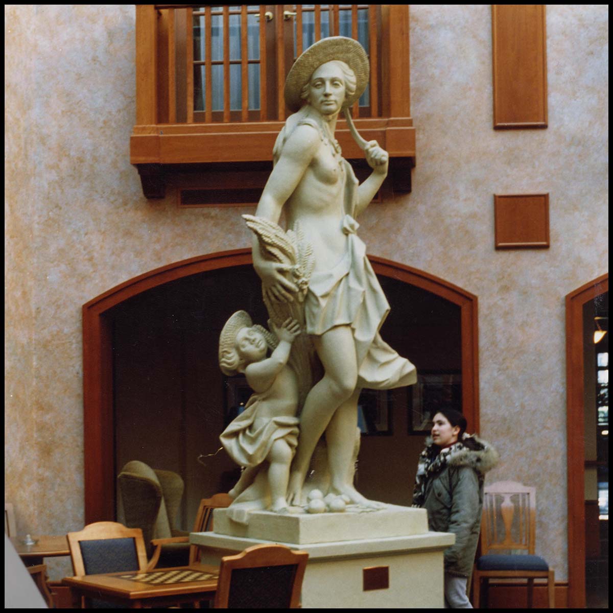 photo of white-colored sculpture of female and child half-clothed and wearing straw hats atop white-colored pedestal in room with blue carpet and wooden tables, chairs, and wood-trimmed doorways with young girl walking behind