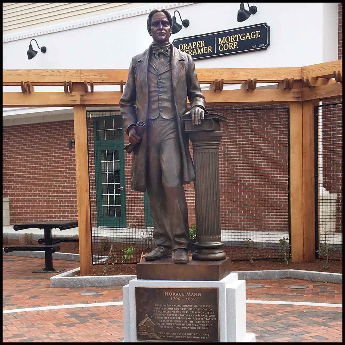 photo of bronze-colored statue of Horace Mann holding scroll and standing beside column with books, atop granite pedestal with bronze-colored plaque in a hardscaped plaza with building behind