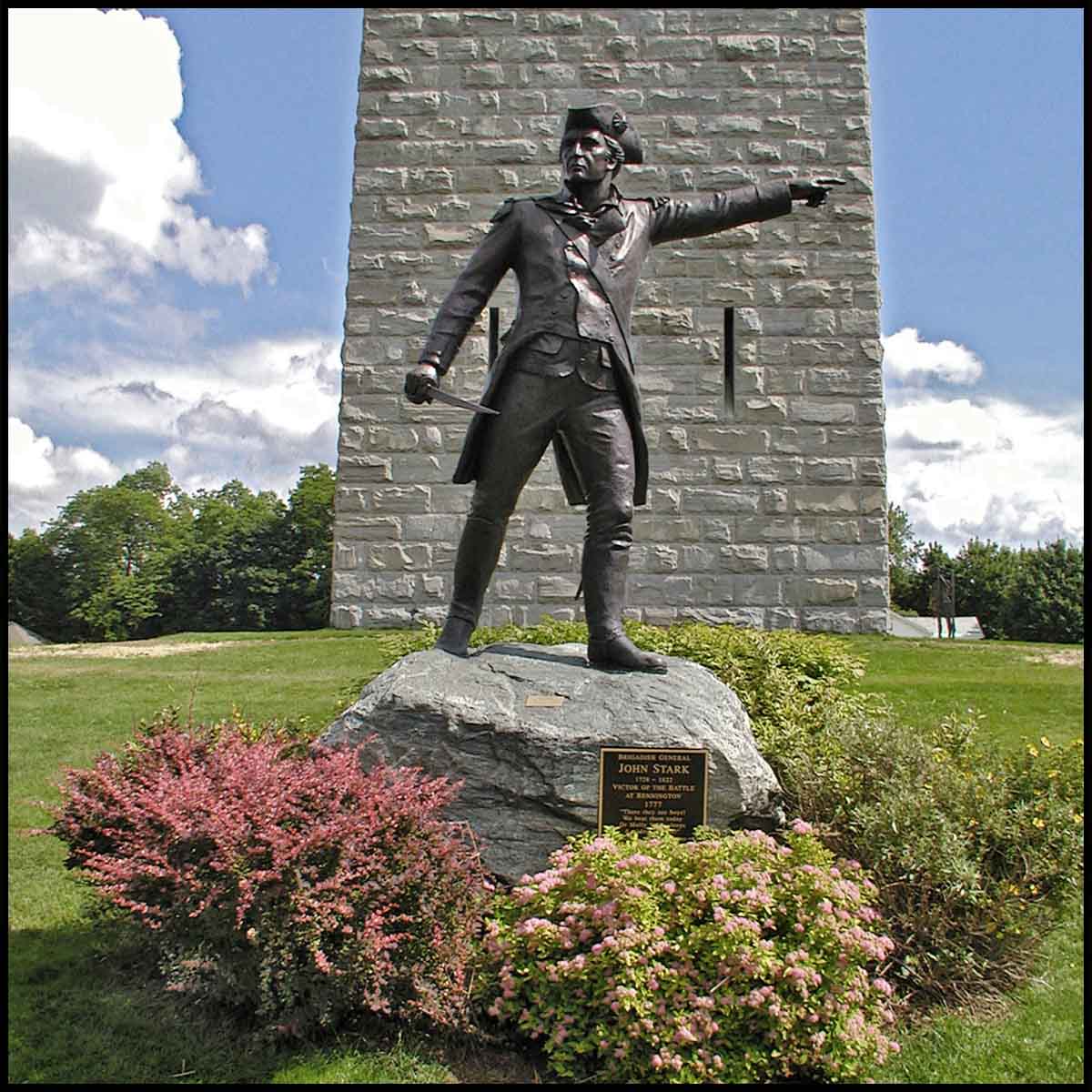 exterior photo of bronze sculpture of John Stark pointing with outstretched arm and standing on rock surrounded by shrubs in front of a obelisk-type monument