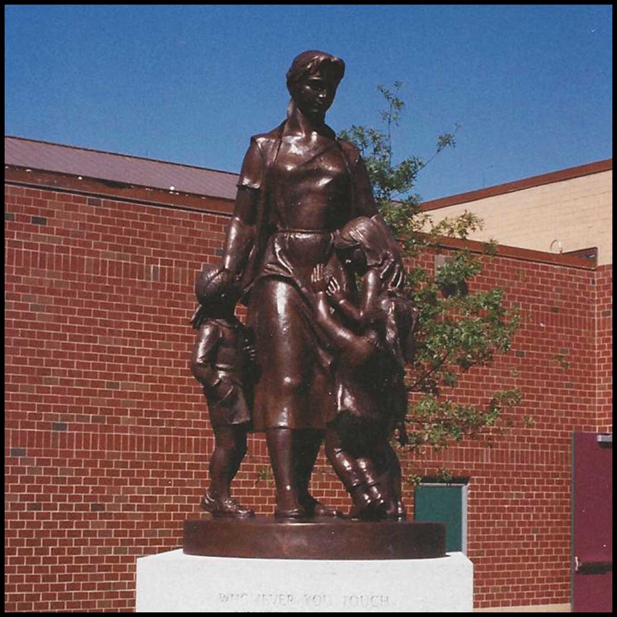 photo of bronze-colored sculpture of female teacher with three children atop granite pedestal on grass in front of brick building