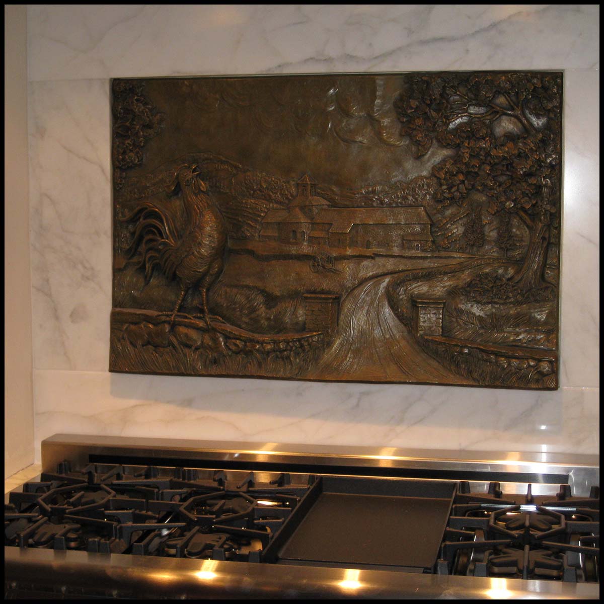 photo of bronze-colored relief plaque of farmhouse with rooster and tree in foreground mounted on tiled wall above kitchen stove