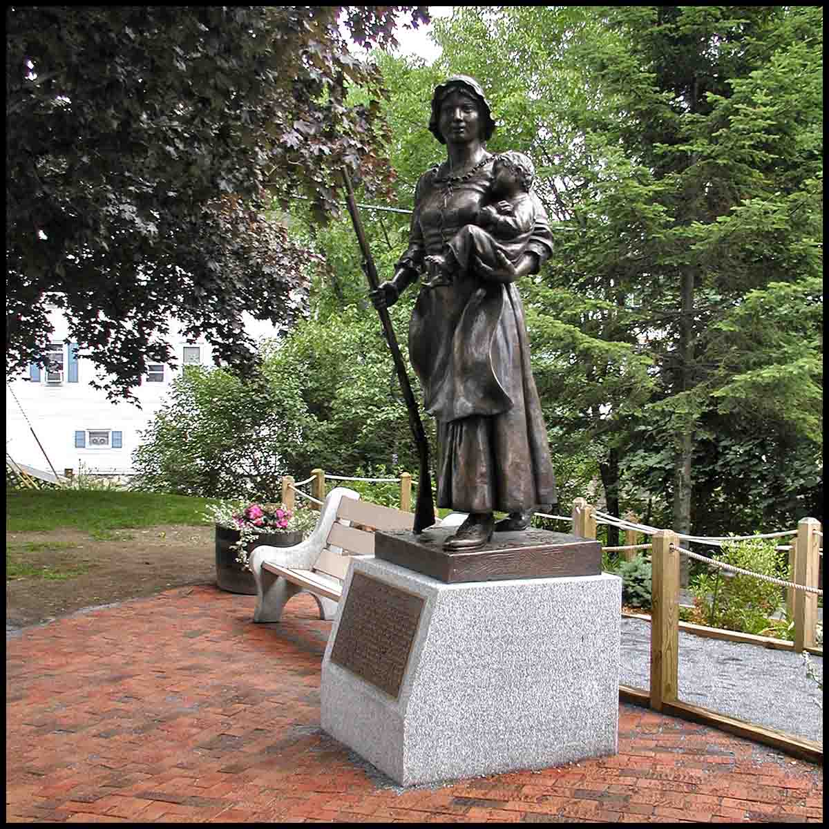 photo of bronze statue of Molly Stark holding child and musket atop stone base with plaque on a brick plaza with surrounding trees