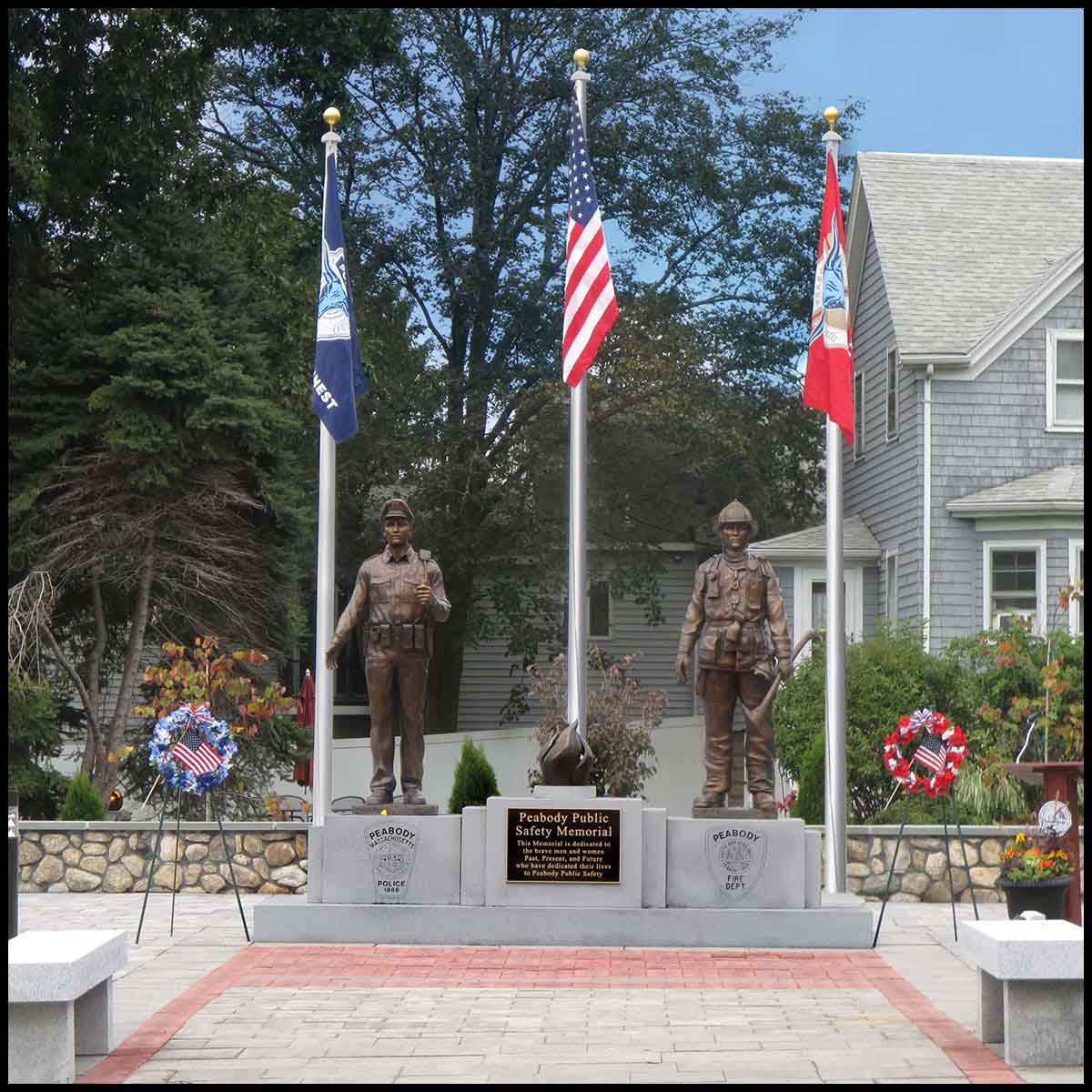 photo of outdoor memorial of bronze sculptures of a firefighter, a police officer, and an eternal flame on a large stone base with etchings and a bronze plaque in a paved site with three flags behind and trees and building behind