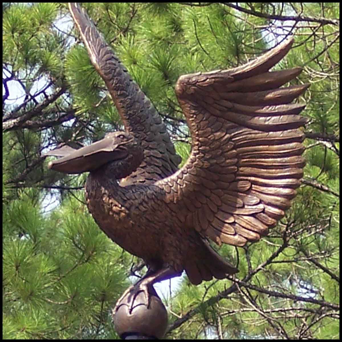 photo closeup of mail kiosk topper of a bronze sculpture of a pelican with letter in beak and surrounded by trees
